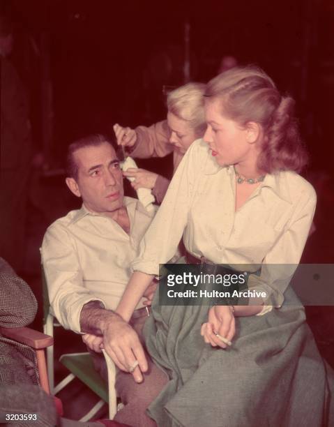 American actor Lauren Bacall sits in the lap of her husband, actor Humphrey Bogart , looking over her shoulder while a makeup artist touches up...
