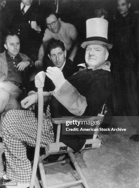 American comedian W.C. Fields holds a wooden cane while sitting in a folding chair in front of crewmembers on the set of director A. Edward...