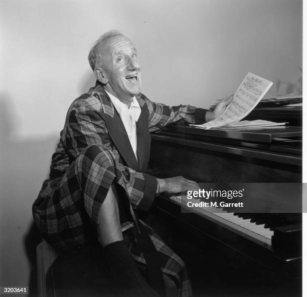 American actor Jimmy Durante wearing a plaid robe at home, seated at a grand piano with one knee elevated, singing and holding sheet music, Los...