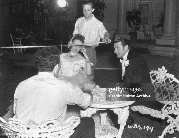 American actors Bette Davis and George Brent sit on lawn furniture while a crew member uses a measuring tape between takes on the set of director...