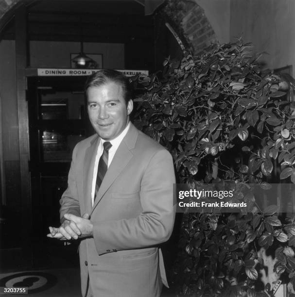 Canadian-born actor William Shatner, wearing a light-colored suit and striped tie, clasps his hands and smiles while standing in front of a shrub...