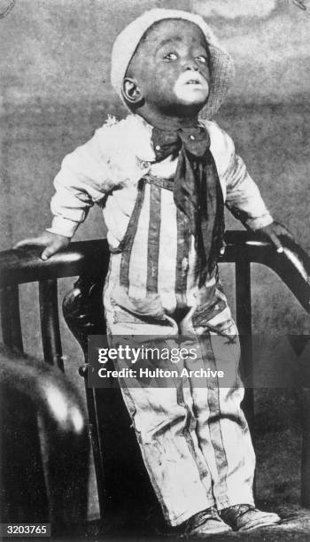 Full-length portrait of American singer and actor Sammy Davis Jr. Posing on a chair in white face makeup, striped overalls, and a cap turned...