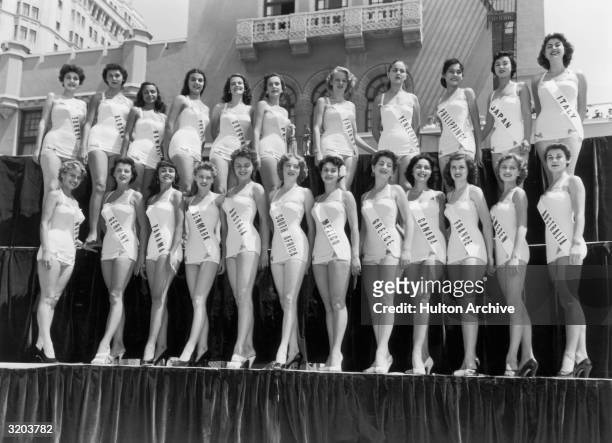 Full-length group portrait of Miss Universe contestants, dressed in bathing suits, smiling while standing on a platform in two rows, Long Beach,...