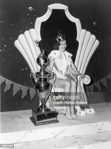 Portrait of Christiane Martel, Miss Universe 1953, sitting on a throne and smiling while holding a scepter next to her trophy, Long Beach,...