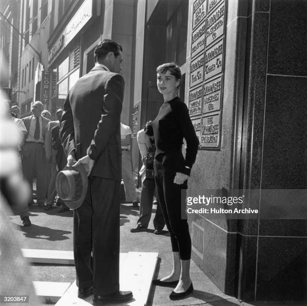 Full-length image of American actor William Holden and Belgian-born actor Audrey Hepburn talking while standing in front of an office building, on...