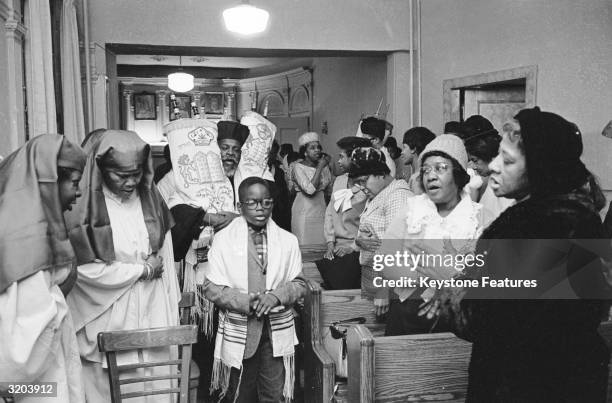 Young Jewish boy leads Rabbi Arthur Matthew, carrying the Torah scrolls, past the congregation in the synagogue on 123rd street in Harlem, New York.