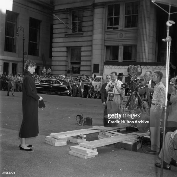 Full-length image of Belgian-born actor Audrey Hepburn putting on gloves while standing on a sidewalk in front of a camera crew, prior to shooting on...