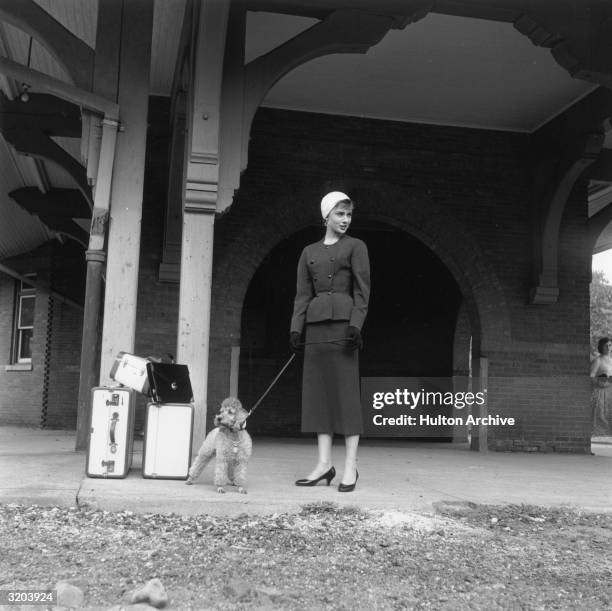 Full-length image of Belgian-born actor Audrey Hepburn waiting in front of a train station with a toy poodle and luggage, on the set of director...