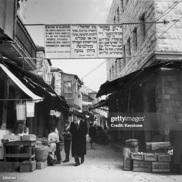 Hassidic Jew walking through the market in Mea Shearim, Jerusalem. Above him hangs a sign commissioned by the Committee for Guarding Modesty.