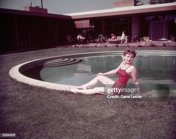 American actor Coleen Gray, wearing a red swimsuit, poses at the edge of a paddock pool outdoors at the home of interior decorator Paul Lazslo,...