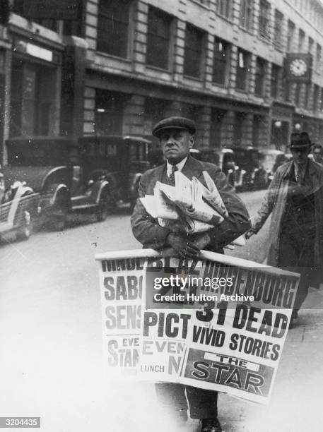 Bill, a London news vendor, carries posters promoting coverage of the 'Hindenburg' disaster in local newspapers, Fleet Street, London, England. He...