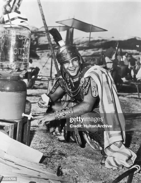 American actor Marlon Brando laughs while crouching next to a water cooler with a cone-shaped paper cup in his hand on the set of director Joseph L....