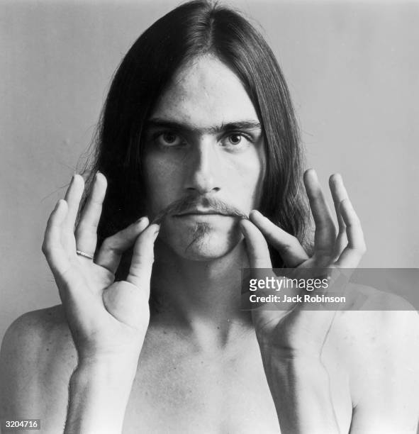 Headshot portrait of American folk musician James Taylor holding the ends of his mustache. He is bare chested.