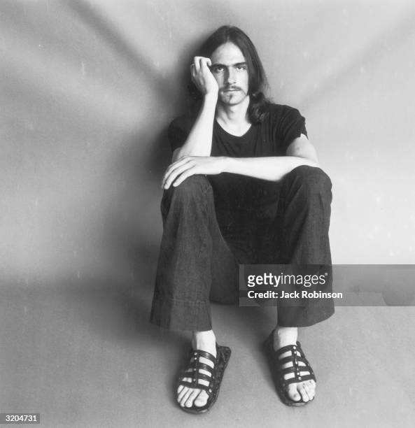 Portrait of American folk musician James Taylor sitting on the floor with his knees pulled up to his chest and his head resting on his hand. He wears...
