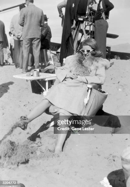 American actor Bette Davis , sitting in a director's chair in the desert, holds a cigarette and plays in the sand with her foot during a break on the...