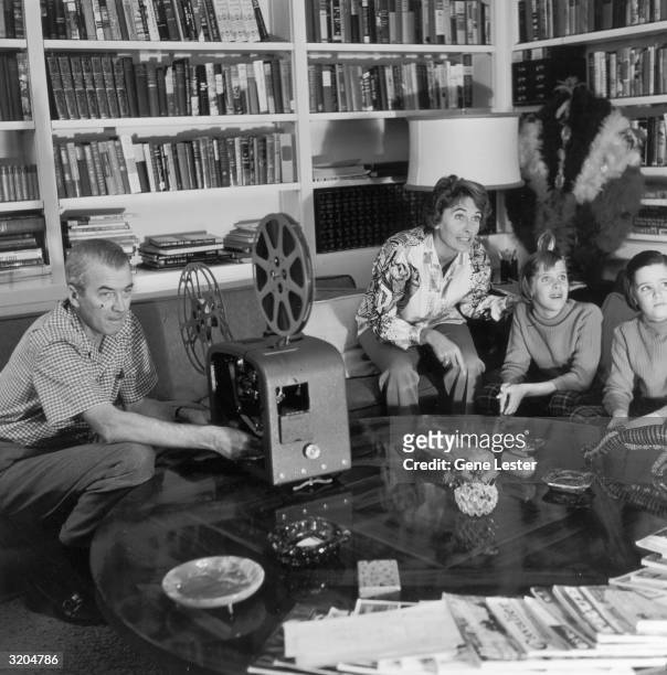 American actor James Stewart operates a film projector while his wife, Gloria, and their twin daughters, Judy and Kelly, watch an unseen movie screen...