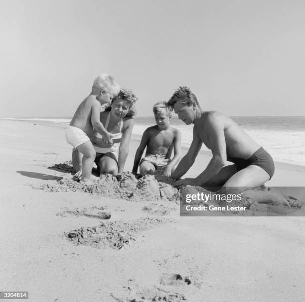 American actor Lloyd Bridges and his wife, Dorothy Simpson, play in the sand with their young sons, Jeff and Beau, on a beach. Each wears a bathing...