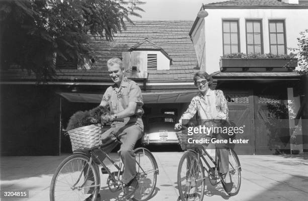 American actor James Stewart and his wife, Gloria Hatrick McLean, ride bicycles in the driveway of their home in California. Their pet poodle sits in...