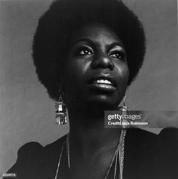 Headshot portrait of American vocalist Nina Simone looking up. She is wearing dangling earrings and a dark top.