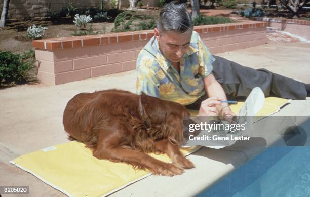 American writer and illustrator Dr Seuss reclines with his Irish setter Cluny and some proofs of his work by the edge of a swimming pool at home in...