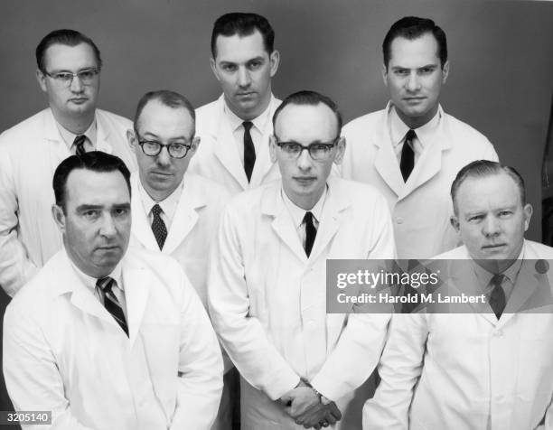 Studio portrait of seven men in white lab coats. Three of the men wear horn-rimmed glasses.