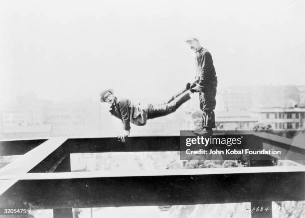 Silent comic Harold Lloyd balances on a high girder while another man holds his legs, in a scene from one of his movies.