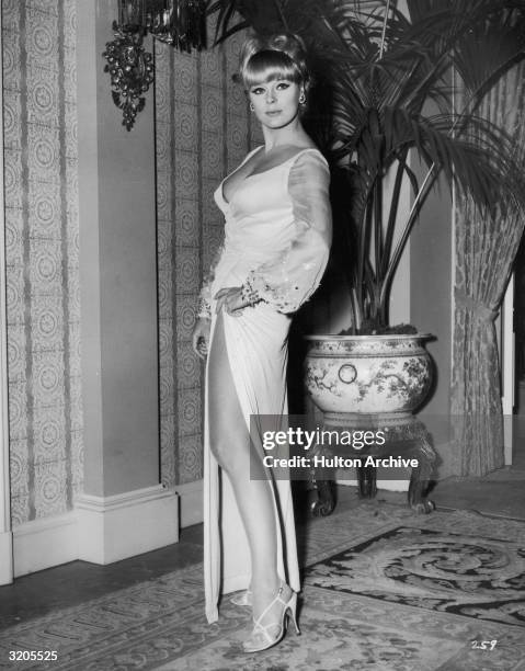 Full-length portrait of German actor Elke Sommer wearing a long gown with a high leg slit and a plunging neckline.
