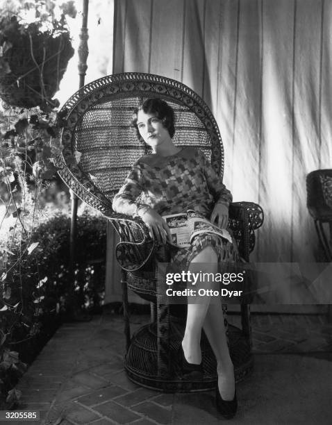 Hollywood film star, Fay Wray, reading a magazine in a wicker chair.