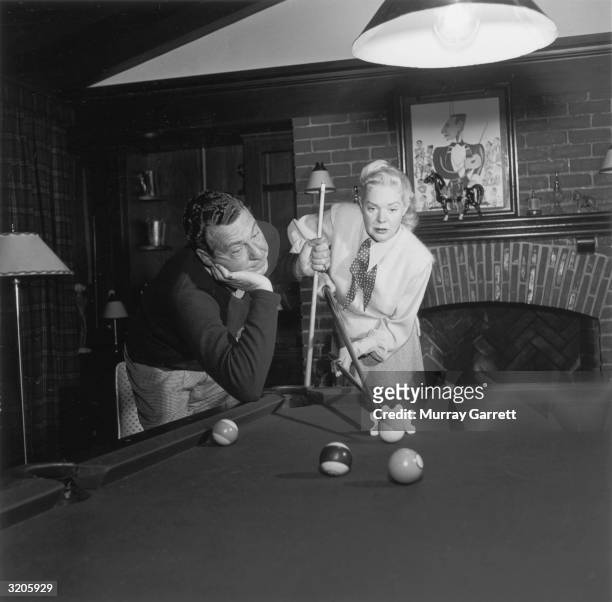American actor and bandleader Phil Harris leans on his elbow, watching his wife, actor and singer Alice Faye, as she lines up a pool shot using the...