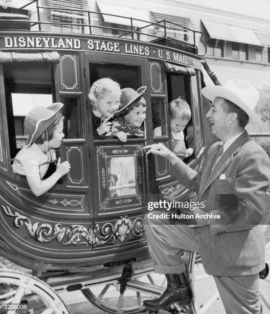 American cartoonist and producer Walt Disney , using a toy revolver, smiles while pretending to hold up children in a stagecoach at his Disneyland...