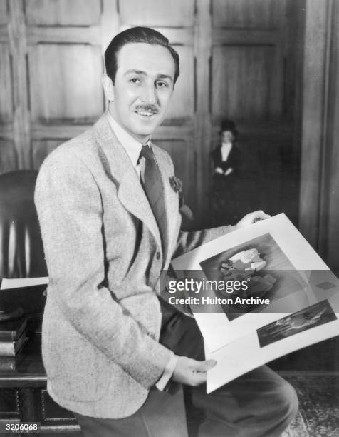 Portrait of American cartoonist and producer Walt Disney seated on the edge of a desk in an office holding illustrations from his animated films...