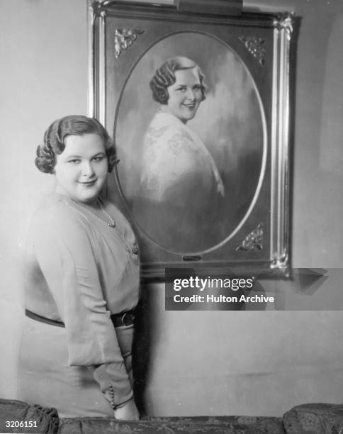 Portrait of American vocalist Kate Smith standing next to a painting of herself and smiling.