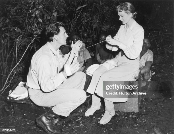 American actor Stewart Granger squats with yarn wrapped around his hands while Scottish actor Deborah Kerr knits, on the set of directors Compton...