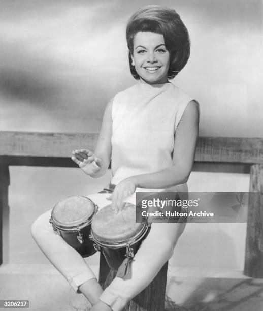 American actor and singer Annette Funicello smiles while playing bongos on a wooden post in a promotional portrait. Funicello has a bouffant and...