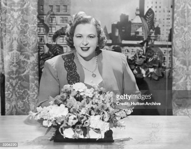 Portrait of American vocalist Kate Smith seated at a table behind a bouquet of flowers. Smith wears a dress suit and a pendant around her neck.