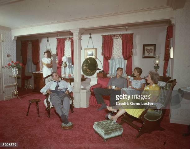American cartoonist and film producer Walt Disney sits with his family while they read and drink tea or coffee in a turn-of-the-century interior,...