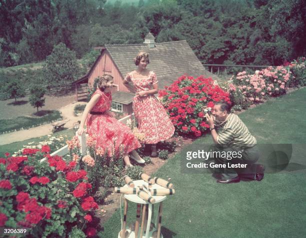 Full-length image of American animator Walt Disney kneeling to take a photograph of his daughters, Sharon and Diane, while they pose in a flower...