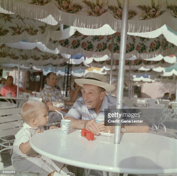 American cartoonist Walt Disney sits with his grandson at an outdoor patio table while visiting Disneyland in Anaheim, California. Both drink from...
