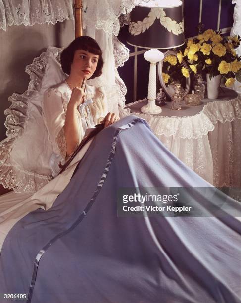 Woman sits in a bed made with white lace sheets and thinks as she writes. A lamp, a mirror, several small bottles, and a vase of yellow flowers sit...
