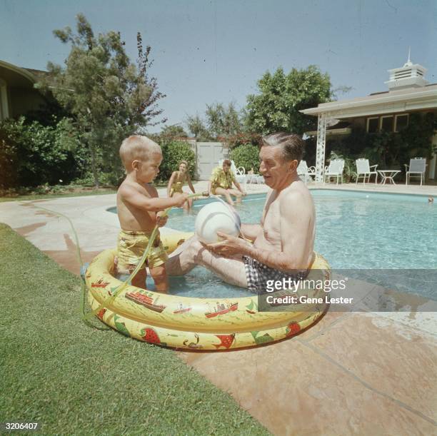 American animator and film studio founder Walt Disney sits and plays with his grandson in a child's inflatable pool, next to a larger swimming pool....