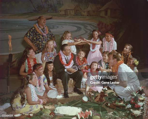 American cartoonist and film producer Walt Disney sings and plays a ukulele for a group of children during a luau-style picnic in front of a tropical...