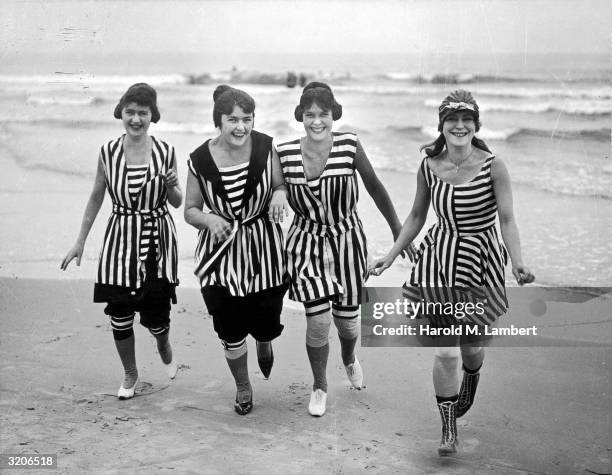 Full-length image of four women laughing while running on a beach in different striped bathing costumes, tights, and lace-up boots or shoes.