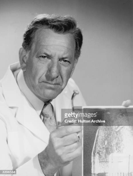 American actor Jack Klugman holds a bullet in a pair of tweezers and an x-ray in a promotional portrait for the television series 'Quincy'. He wears...