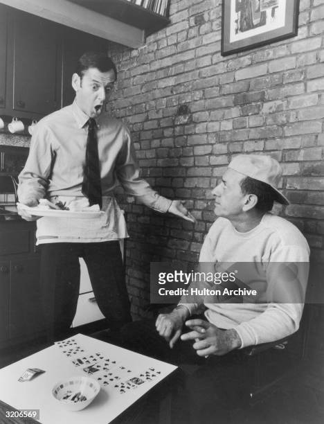 American actor Tony Randall holds a tray of snacks while shouting at American actor Jack Klugman, who plays solitaire while holding a cigar, in a...