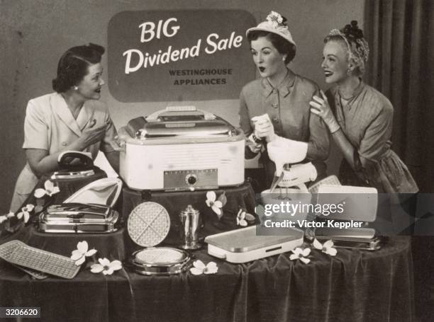 Salesperson shows an array of Westinghouse electric kitchen appliances, including a waffle-iron, egg-beater and iron, to two women who look...