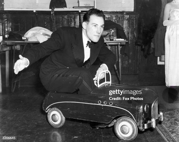 British racing driver, Stirling Moss arrives driving a toy car at the annual masked ball held by the Enfield Arts Circle, where he is to judge the...