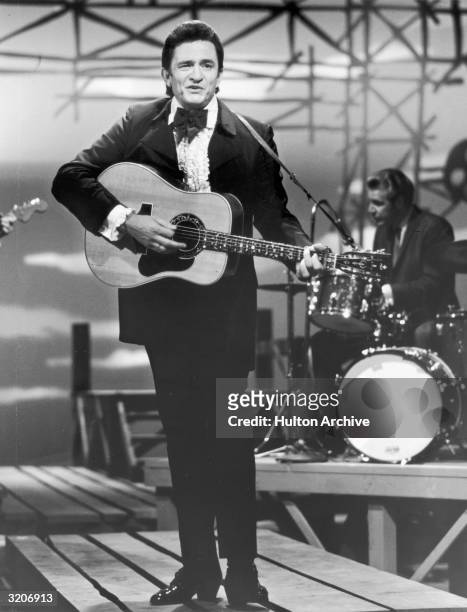 Full-length image of American country singer and musician Johnny Cash , wearing a tuxedo, performing with a guitar on stage during a television...