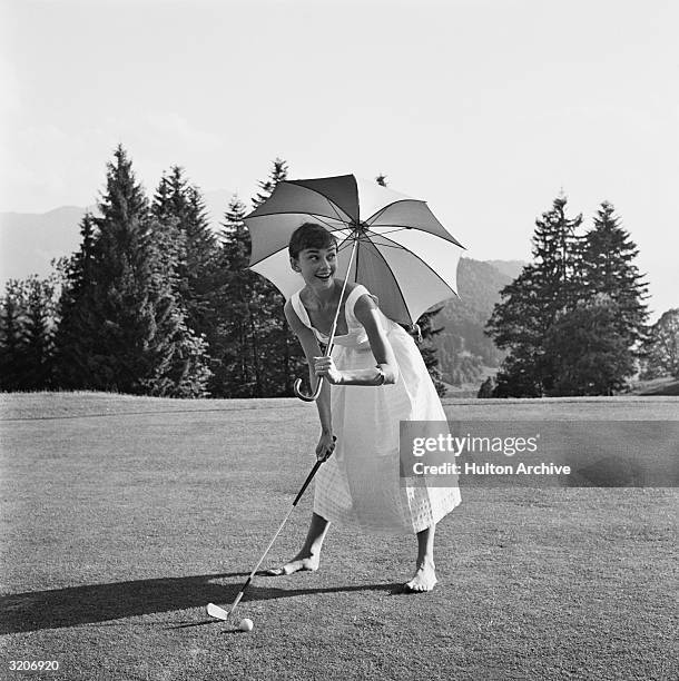 Belgian-born actor Audrey Hepburn , barefoot and wearing a summer dress, playing golf and holding an umbrella over her head.