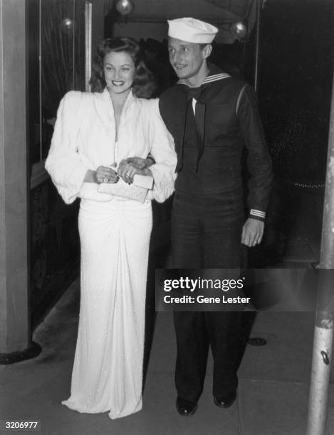 Full-length image of American actor Gene Tierney being escorted by her husband, French fashion designer Oleg Cassini, who wears a naval uniform. She...