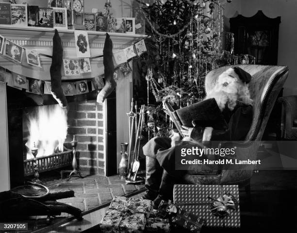 Man dressed as Santa Claus sits in an armchair in a den, reading a large book by the fireplace. There is a decorated Christmas tree and wrapped...